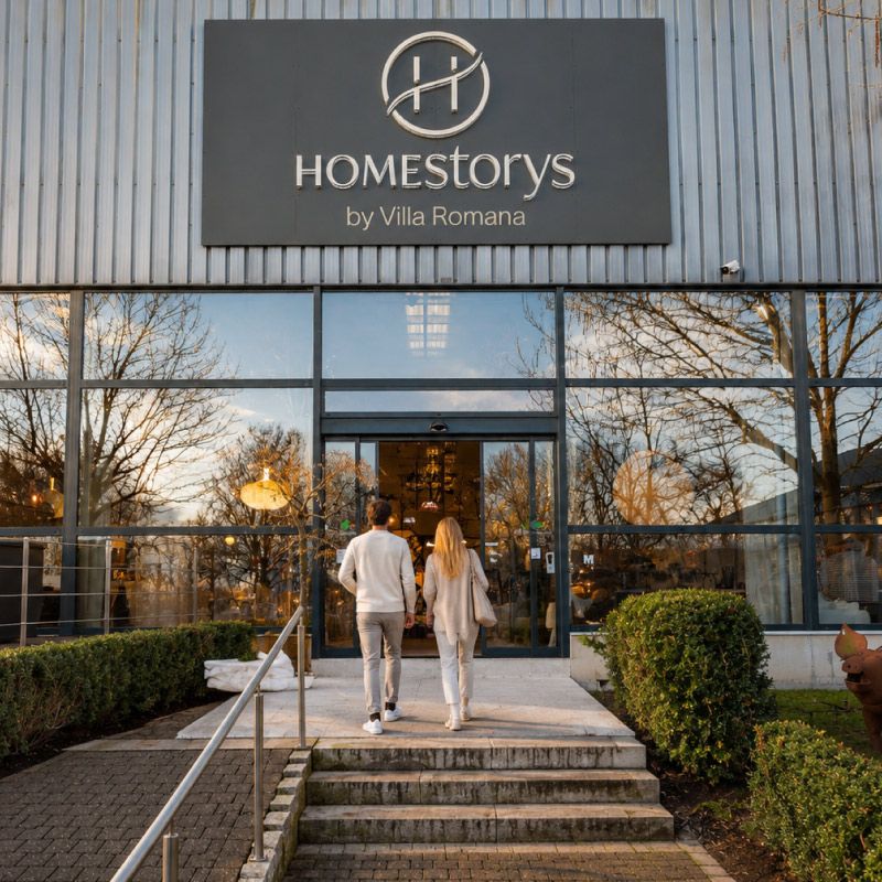 Homestorys zieht um – Eingang unseres Showrooms Ein Paar betritt den Homestorys-Showroom; über dem Eingang ist das vollständige Homestorys-Logo zu sehen.